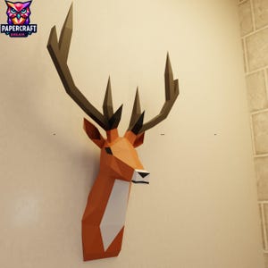May include: A geometric papercraft deer head in shades of orange, brown, and white, mounted on a beige wall. The antlers are dark brown, and the head features a white chest. The Papercraft Dream logo is in the upper left corner.