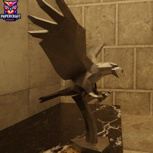May include: A metallic, geometric eagle sculpture with outstretched wings, perched on a marble-like base. The sculpture has a polygonal design and is set against a textured wall. The Papercraft Dream logo is in the upper left corner.