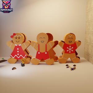 May include: Three gingerbread man decorations with smiling faces and various outfits. One wears a red dress, another red overalls, and the third a red shirt with a white snowflake. The Papercraft Dream logo is in the upper left corner.