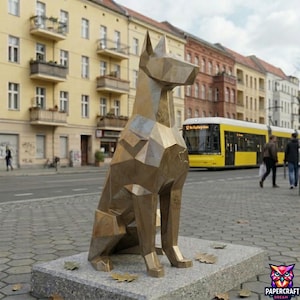 Könnte beinhalten: Eine geometrische Hundeskulptur aus Bronze sitzt auf einem Steinsockel. Der Hund sitzt mit aufgestellten Ohren. Die Skulptur befindet sich im öffentlichen Raum mit Gebäuden und einem gelben Bus im Hintergrund.