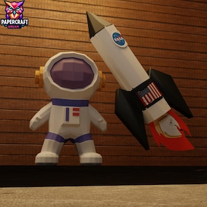 Op de afbeelding: Een witte en paarse astronautenfiguur en een witte raket met zwarte vinnen. De raket heeft een NASA-logo en een Amerikaans vlagontwerp. De astronautenfiguur heeft een paars vizier en goudkleurige oorschelpen. De raket heeft rode vlammen.