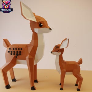 May include: Two papercraft deer figures. The larger deer is brown with black spots and white accents. The smaller deer is also brown with white accents. The figures have geometric designs and are standing on a yellow surface. The logo "PAPERCRAFT DREAM" is in the top left corner.