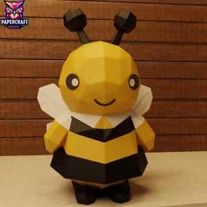 Puede incluir: Una figura geométrica de abeja de papel con rayas amarillas y negras, alas blancas y antenas negras. La abeja tiene una cara sonriente y está sobre un fondo de madera. El logotipo "PAPERCRAFT DREAM" es visible en la esquina superior izquierda.