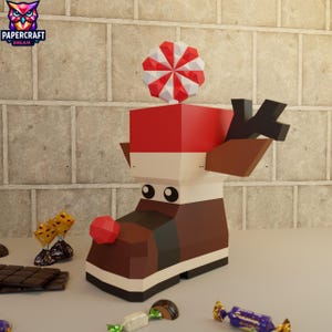 Op de afbeelding: Een grillige papieren rendierlaars met een rood-witte pepermunt op de top. De laars is bruin, wit en rood, met een rode neus en zwarte geweien. Verschillende snoepjes en een chocoladereep liggen rond de laars.