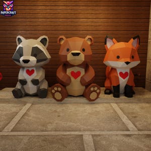 Puede incluir: Tres figuras geométricas de animales de papel: un mapache, un oso y un zorro. Cada figura tiene un corazón rojo en el pecho. El mapache es gris y blanco, el oso es marrón y el zorro es naranja y blanco. Las figuras están sobre una superficie de piedra.