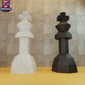 May include: Two large, geometric chess king pieces. One is white with a cross on top, the other is black. The pieces are on a yellow surface with a textured, light brown background. The logo "PAPERCRAFT DREAM" is in the upper left corner.