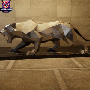 Puede incluir: Una escultura geométrica plateada de una pantera rugiente. La obra de arte está hecha de paneles metálicos angulares, lo que le da una apariencia moderna y facetada. La escultura se exhibe sobre una base oscura y rectangular, con el logotipo de Papercraft Dream en la esquina superior izquierda.
