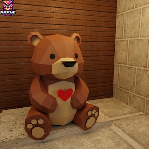 May include: A brown papercraft bear with a red heart on its chest. The bear has a geometric design and is sitting down. The bear has black eyes and a black nose. The bear is sitting on a stone surface.