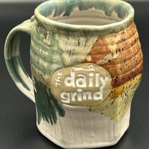 May include: A ceramic mug with a textured, multi-colored glaze featuring shades of green, orange, yellow, and white. An oval-shaped medallion on the front displays the embossed white text "THE daily grind" against a tan background.