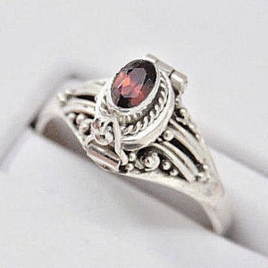 May include: A silver ring with a red oval gemstone set in a detailed filigree design. The ring has a hinged opening, revealing a small compartment inside.
