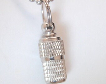 May include: A silver metal necklace with a small, cylindrical pendant. The pendant has a textured surface and a small loop at the top.