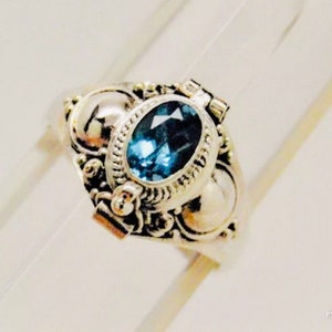 May include: A silver ring with an oval, dark blue gemstone at the center. The ring has an ornate, detailed design with decorative elements around the gemstone and on the band. The ring is set against a light background.