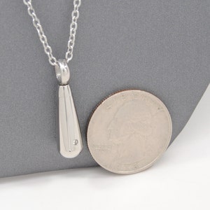 May include: A silver pendant necklace with a teardrop-shaped charm. The charm has a small diamond-shaped stone at the bottom. The necklace is shown next to a US quarter for size comparison.