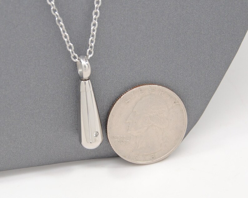 May include: A silver pendant necklace with a teardrop-shaped charm. The charm has a small diamond-shaped stone at the bottom. The necklace is shown next to a US quarter for size comparison.