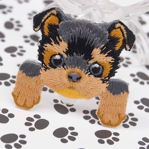 May include: Embroidered patch of a Yorkshire Terrier puppy with brown, black, and gray fur. The puppy is looking at the viewer with its paws outstretched.
