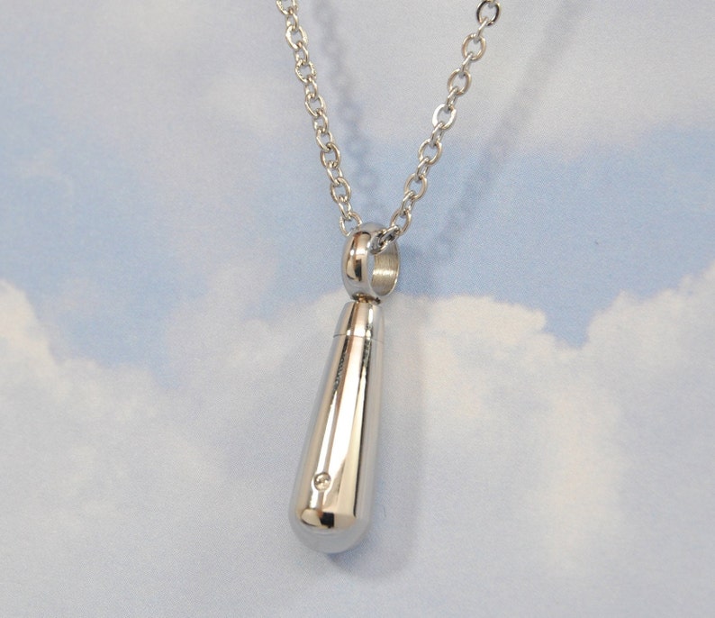 May include: A silver teardrop-shaped pendant necklace with a small, round opening at the top. The pendant is attached to a silver chain.