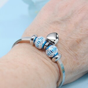 May include: A silver bangle bracelet with a heart charm and two blue and silver patterned beads.