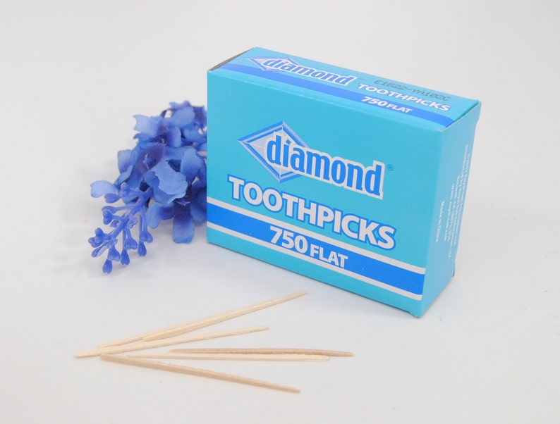 May include: A blue box of Diamond toothpicks with 750 flat toothpicks inside. The box is on a white surface with a small bouquet of blue flowers.
