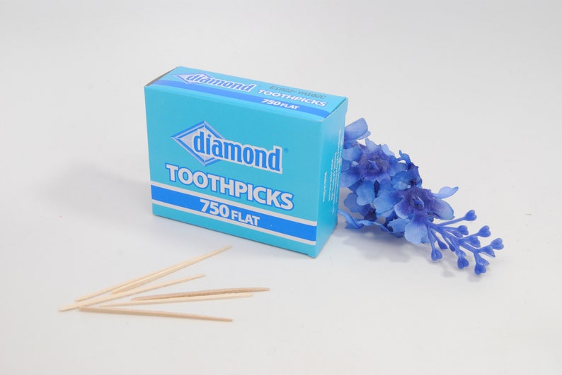 May include: A blue box of Diamond toothpicks with 750 flat toothpicks. The box is on a white surface with a few toothpicks scattered in front of it.