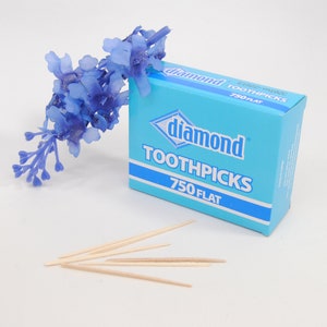 May include: A blue box of Diamond toothpicks with 750 flat toothpicks. The box is sitting on a white surface with a small bouquet of blue flowers.