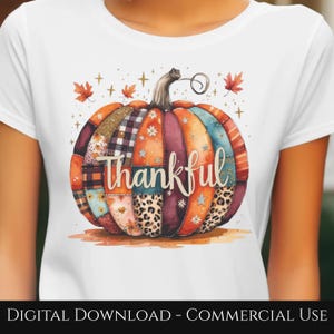 May include: White t-shirt featuring a watercolor pumpkin design. The pumpkin is composed of various patterned sections in orange, brown, and blue, with the word "thankful" in script. Autumn-themed with leaves and stars.