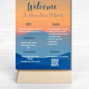 May include: A welcome sign for a mountain retreat with a blue and orange mountain background. The sign includes information about wifi, address, contacts, rules, and a review QR code. The text on the sign reads: "Welcome to Mountain Retreat", "Wifi", "Network: Spectrum 92-9", "Password: inthemountains", "Address", "123 Mountain Drive", "Mountain Town, NC", "Contacts", "Host: 555-555-5555", "Email: host@mountains.com", "Rules", "• No Smoking or Parties", "• Take trash out and roll can to the road on checkout day", "• Put A/C at 75 at checkout", "• Have Fun!", "Review", "Scan to leave us a review!"