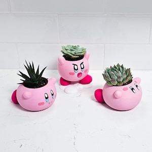 May include: Three pink ceramic succulent planters shaped like the video game character Kirby. Each planter has a different facial expression and features a small green succulent plant.