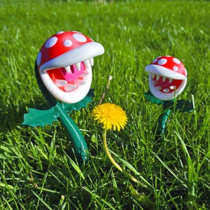 May include: Two Piranha Plant figures from the Super Mario video game franchise. The plants have red and white spotted heads, white teeth, and pink tongues. They are set in green grass with a yellow dandelion.