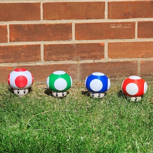 May include: Four colorful mushroom figurines with white spots and stems, arranged on green grass in front of a brick wall. The mushrooms have red, green, blue, and red caps.