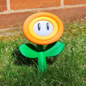 May include: A 3D-printed, orange and yellow flower with a white center and black oval eyes, resembling a character from a video game. The flower has green leaves and stem, standing in grass. The flower is a decorative item.