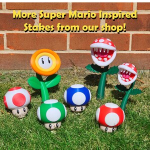 May include: A selection of Super Mario-themed garden stakes, featuring a yellow flower, red and white mushrooms, a green mushroom, a blue mushroom, and two Piranha Plants. The stakes are displayed on grass in front of a brick wall. The text reads "More Super Mario Inspired Stakes from our shop!"