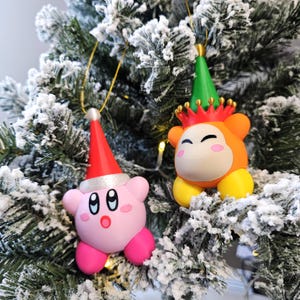 May include: Two Christmas tree ornaments. One is pink with a red Santa hat and the other is orange with a green party hat and a crown. Both ornaments have gold hanging strings and are on a snowy green Christmas tree.