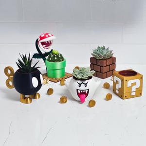 May include: Five different plant pots shaped like characters from the video game Super Mario Bros. The pots are white, green, black, brown, and gold. Each pot has a different plant growing in it. The pots are sitting on a white surface.