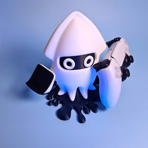 Op de afbeelding: Een witte inktvisachtige figuur met zwarte accenten, die een wit-zwarte gamecontroller vasthoudt. De figuur staat op een zwarte, gespatte basis tegen een lichtblauwe achtergrond. De controller heeft witte knoppen en een zwart touchpad.