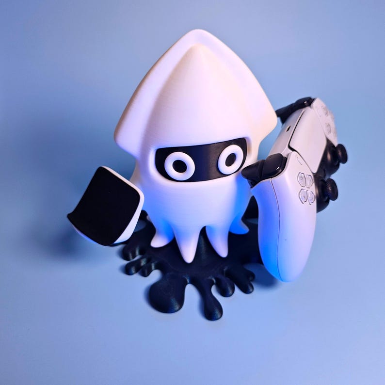 Op de afbeelding: Een witte inktvisachtige figuur met zwarte accenten, die een wit-zwarte gamecontroller vasthoudt. De figuur staat op een zwarte, gespatte basis tegen een lichtblauwe achtergrond. De controller heeft witte knoppen en een zwart touchpad.