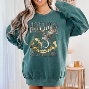 May include: Teal-colored sweatshirt with a graphic design featuring a silver dragon, a gold dragon, and the text "ONYX STORM." Below the dragons, a banner reads "THE BIGGEST BATTLE YET BRAVE THE DARK."