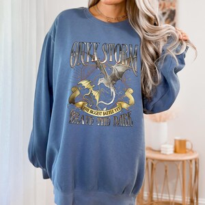 May include: A blue sweatshirt with the text "ONYX STORM" above a graphic of two dragons. The text "THE BIGGEST BATTLE YET" and "BRAVE THE DARK" are below the dragons. The sweatshirt has long sleeves.