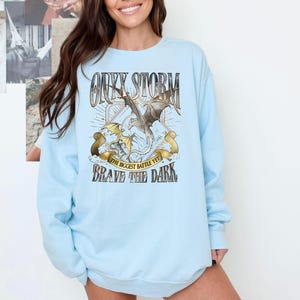 May include: Light blue sweatshirt with a graphic featuring a dragon and the text "Onyx Storm, The Biggest Battle Yet, Brave the Dark." The design includes a dragon with outstretched wings, set against a backdrop of rays and banners.