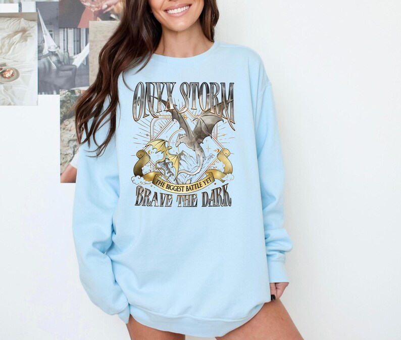 May include: Light blue sweatshirt with a graphic featuring a dragon and the text "Onyx Storm, The Biggest Battle Yet, Brave the Dark." The design includes a dragon with outstretched wings, set against a backdrop of rays and banners.