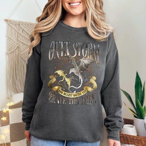 May include: Dark gray sweatshirt with a dragon graphic. The design includes the words "ONYX STORM", "THE BIGGEST BATTLE YET", and "BRAVE THE DARK". The graphic features two dragons in gold and silver.