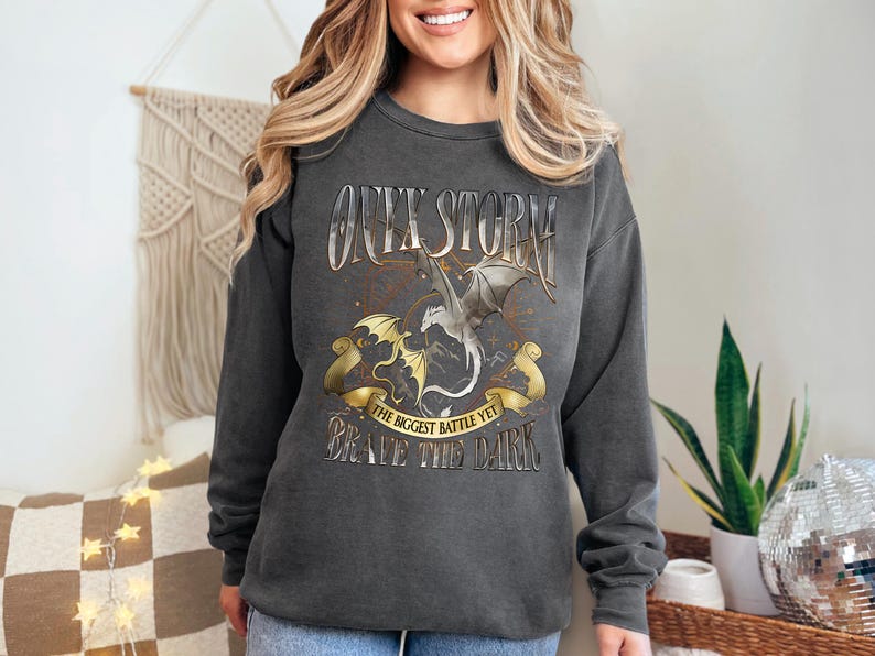 May include: Dark gray sweatshirt with a dragon graphic. The design includes the words "ONYX STORM", "THE BIGGEST BATTLE YET", and "BRAVE THE DARK". The graphic features two dragons in gold and silver.