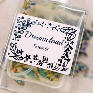May include: A clear glass bottle with a white label that says "Dreamcloud Serenity" in a black floral frame. The bottle is filled with a clear liquid and has a small amount of dried flowers inside.