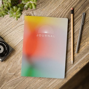 May include: A journal with a colorful gradient cover, featuring yellow, orange, green, and gray hues. The word "JOURNAL" is printed in white. A pencil and pen rest beside the journal on a wooden surface.