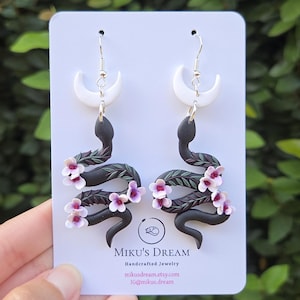 May include: A pair of dangle earrings featuring a black snake coiled around a white crescent moon, adorned with small pink flowers and green leaves. The display card reads MIKU'S DREAM Handcrafted Jewelry and includes the URL mikusdream.etsy.com.
