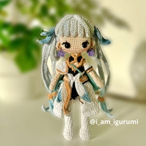 Può includere: Una bambola all'uncinetto con lunghi capelli bianchi, vestita con un abito blu e bianco con una cintura dorata. La bambola ha un cappello all'uncinetto blu chiaro e bianco e orecchini viola. La bambola è in piedi su una superficie bianca. Il testo "@i_am_igurumi" è visibile nell'angolo in basso a destra dell'immagine.