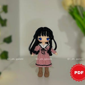 Peut inclure: Poupée au crochet avec des cheveux noirs et des yeux bleus, vêtue d'une robe rose avec un col blanc et des bottes marron. La poupée a de longs cheveux noirs et se tient sur une surface blanche. L'image comprend le texte "PDF" dans un cercle rouge.