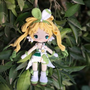 May include: A crocheted doll with yellow hair, green and white clothing, and blue eyes. The doll is wearing a flower crown and is posed with its arms outstretched.