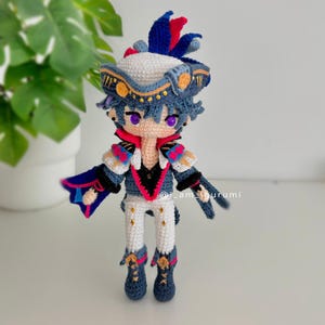 Può includere: Bambola amigurumi all'uncinetto con abiti blu e bianchi, un cappello bianco con bordi dorati e occhi viola. La bambola indossa una giacca blu e bianca con un colletto rosso e rosa. La bambola è in piedi su una superficie bianca.