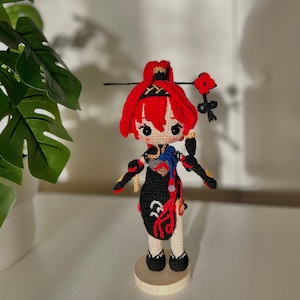 Puede incluir: Una muñeca de crochet con cabello rojo, un vestido negro y una flor roja en la cabeza. La muñeca está de pie sobre una base de madera.