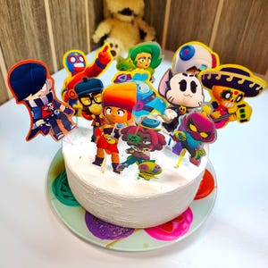 May include: A white cake decorated with colorful cartoon character cake toppers. The cake toppers are of various characters from the video game Brawl Stars.